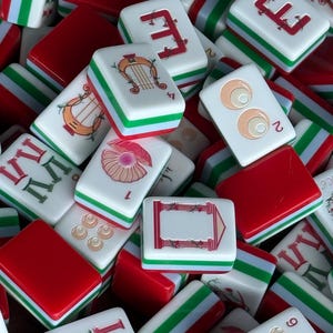 May include: A collection of rectangular Mahjong tiles featuring white faces with red, green, and tan illustrations including a harp, a shell, eyes, and Roman numerals. The tiles have red and green layered sides and are arranged in a scattered pile.