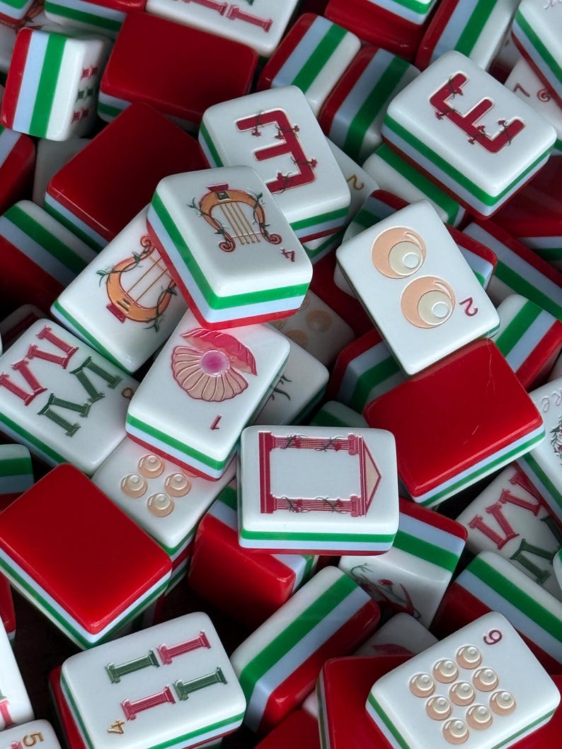 May include: A collection of rectangular Mahjong tiles featuring white faces with red, green, and tan illustrations including a harp, a shell, eyes, and Roman numerals. The tiles have red and green layered sides and are arranged in a scattered pile.
