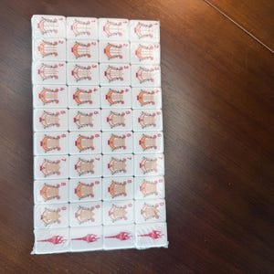 May include: A rectangular set of white mahjong tiles arranged in a grid, featuring orange harp illustrations numbered one through nine in each column, with red torch-like symbols on the bottom row, all encased in clear plastic shrink wrap.