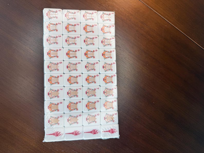 May include: A rectangular set of white mahjong tiles arranged in a grid, featuring orange harp illustrations numbered one through nine in each column, with red torch-like symbols on the bottom row, all encased in clear plastic shrink wrap.