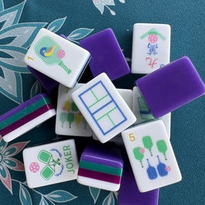 May include: A collection of white Mahjong tiles with various colorful illustrations and numbers. The tiles feature images of rackets, balls, and other game-related symbols. Some tiles have purple and green stripes, and the word "JOKER" is visible on one tile.