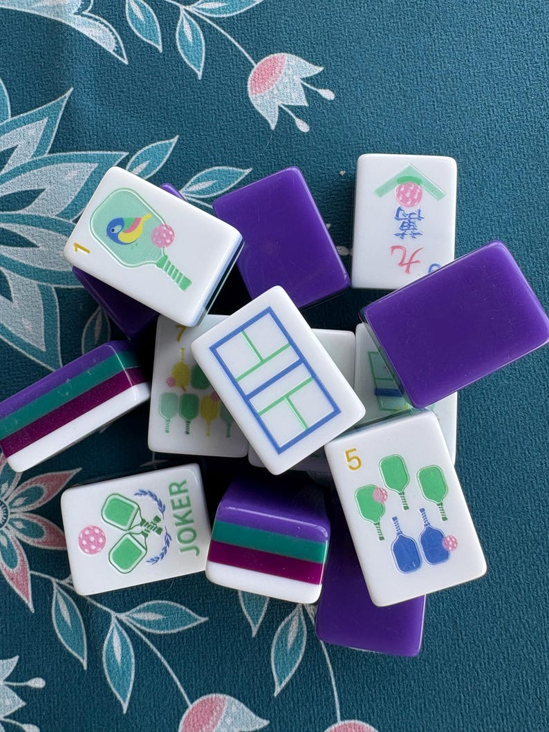 May include: A collection of white Mahjong tiles with various colorful illustrations and numbers. The tiles feature images of rackets, balls, and other game-related symbols. Some tiles have purple and green stripes, and the word "JOKER" is visible on one tile.