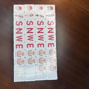 May include: A set of rectangular white mahjong tiles arranged in a grid, featuring red and green floral designs, red lettered directional indicators including S, N, W, and E, and tiles depicting a golden lyre symbol with the word Joker printed below.