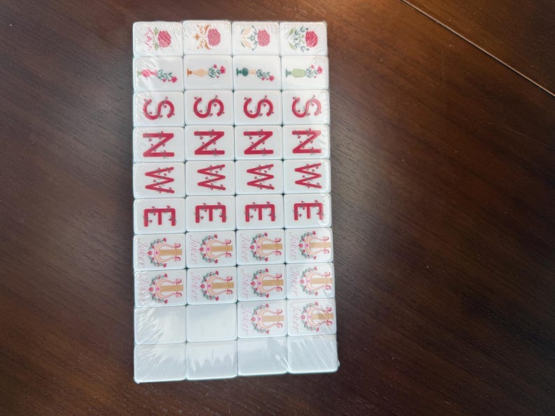 May include: A set of rectangular white mahjong tiles arranged in a grid, featuring red and green floral designs, red lettered directional indicators including S, N, W, and E, and tiles depicting a golden lyre symbol with the word Joker printed below.
