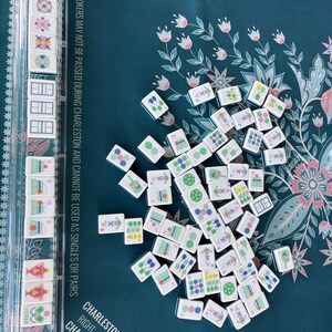 May include: A collection of white Mahjong tiles with various symbols and numbers scattered on a teal floral patterned surface. A clear plastic tile holder is visible on the left. The game board has text that reads "Charleston (Required)".