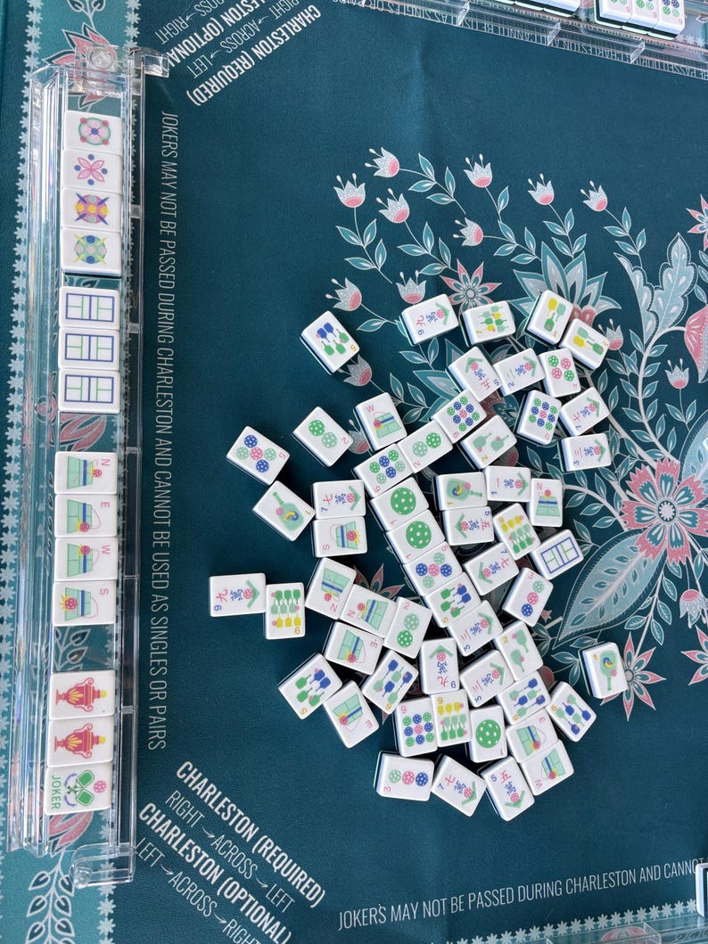 May include: A collection of white Mahjong tiles with various symbols and numbers scattered on a teal floral patterned surface. A clear plastic tile holder is visible on the left. The game board has text that reads "Charleston (Required)".