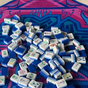May include: A collection of rectangular Mahjong tiles with white faces featuring various green, red, and blue symbols, and solid blue backs. The tiles are scattered across a bright pink and blue game mat printed with text instructions.