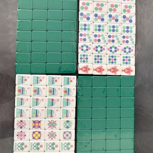 May include: A set of Mahjong tiles, packaged in plastic wrap. The tiles are green and white, with various symbols and numbers. Some tiles have the word "JOKER" printed on them. The game pieces are arranged in four stacks.