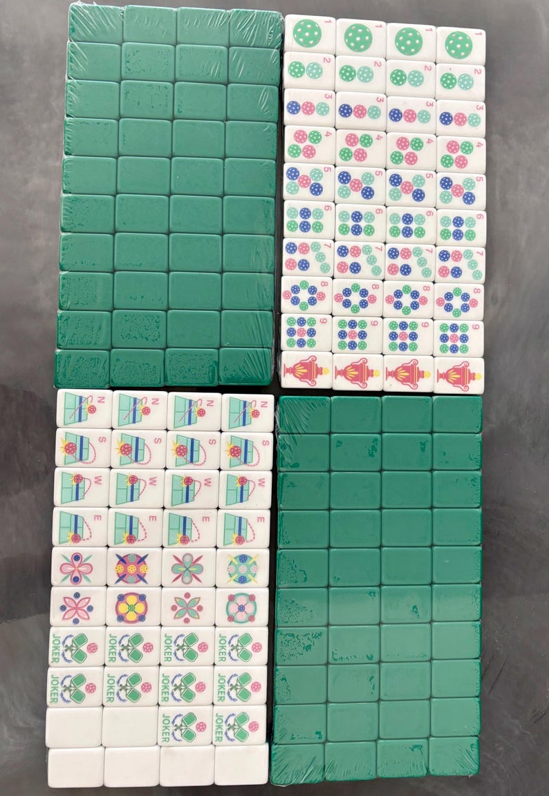 May include: A set of Mahjong tiles, packaged in plastic wrap. The tiles are green and white, with various symbols and numbers. Some tiles have the word "JOKER" printed on them. The game pieces are arranged in four stacks.