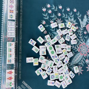 May include: A collection of white Mahjong tiles with various symbols and numbers scattered on a teal floral patterned surface. A clear plastic tile holder is visible on the left. The game board has text that reads "Charleston (Required)".