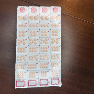 May include: A set of white rectangular mahjong tiles arranged in a grid and wrapped in clear plastic. Each tile features a number from one to nine represented by orange circular icons, with a pink floral design for the one and red frames at the bottom.