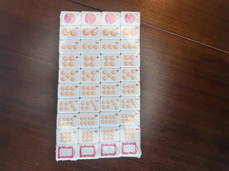 May include: A set of white rectangular mahjong tiles arranged in a grid and wrapped in clear plastic. Each tile features a number from one to nine represented by orange circular icons, with a pink floral design for the one and red frames at the bottom.