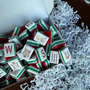May include: A collection of rectangular mahjong tiles with red, white, and green edges inside an open white cardboard box. The tiles feature various printed designs including the letter W, a harp, the letter S, and numerical symbols in red and green.