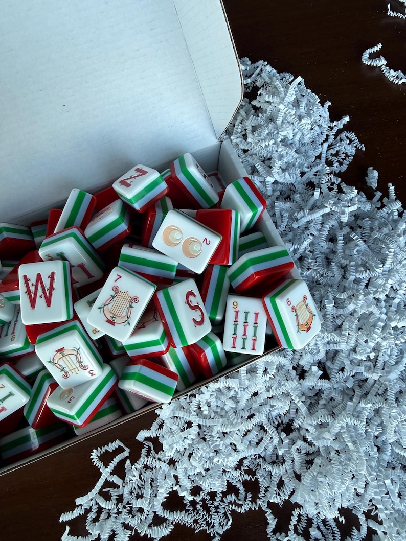 May include: A collection of rectangular mahjong tiles with red, white, and green edges inside an open white cardboard box. The tiles feature various printed designs including the letter W, a harp, the letter S, and numerical symbols in red and green.