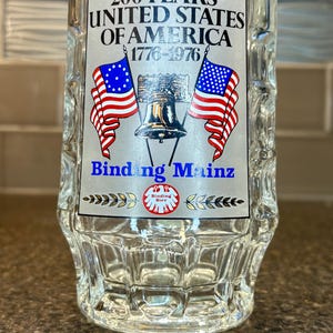 May include: Clear glass beer stein with a decorative label commemorating the 200th anniversary of the United States of America. The label features the text "200 Years United States of America 1776-1976" with illustrations of American flags and the Liberty Bell.