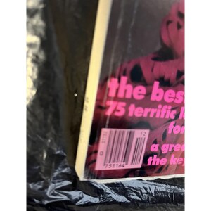 Puede incluir: Una portada de libro con el texto rosa "the bes 75 terrific" sobre un fondo oscuro. Un c&oacute;digo de barras y texto adicional son visibles en la esquina inferior derecha. El libro est&aacute; envuelto en pl&aacute;stico transparente.
