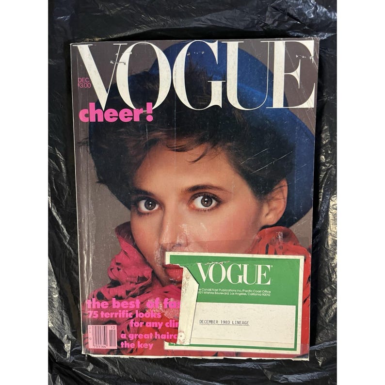 Puede incluir: Una portada de la revista Vogue vintage con una mujer de pelo oscuro y un sombrero azul. El t&iacute;tulo de la revista "VOGUE" se muestra de forma destacada, con la palabra "cheer!" en rosa. La portada incluye texto sobre moda y peinados.