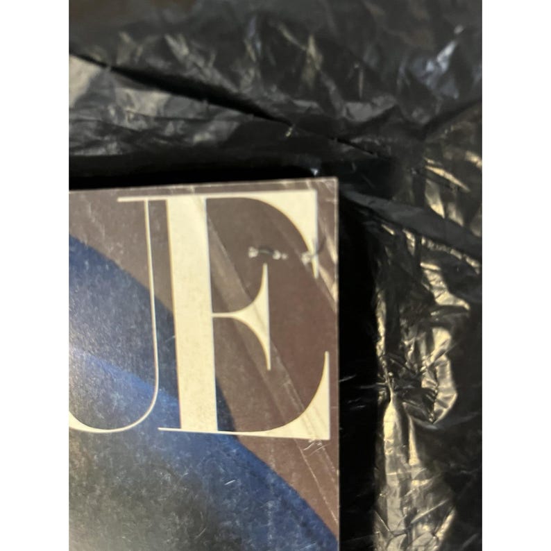 Puede incluir: Primer plano de una portada de revista con la palabra "VOGUE" en fuente grande, blanca y serif. El fondo presenta formas abstractas azules y marrones. La revista est&aacute; parcialmente oscurecida por una l&aacute;mina de pl&aacute;stico negro.