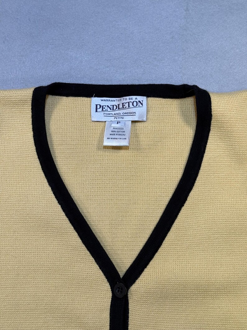 May include: A yellow cardigan with black trim and a V-neck. The cardigan has a single black button closure and a tag that reads "Pendleton, Portland, Oregon". The cardigan is made of a knit material.