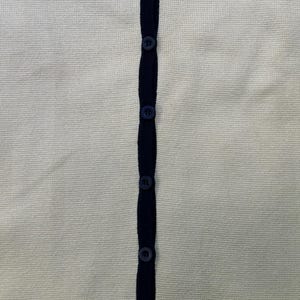May include: Cream-colored cardigan with navy blue trim and buttons. The button-up sweater features a V-neckline and a classic, preppy style. The knitwear is a versatile piece for layering.
