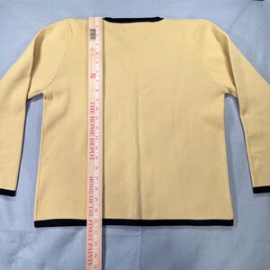 May include: A light yellow sweater with long sleeves and a crew neck. The sweater has black trim on the neckline, cuffs, and hem. A Home Depot ruler is placed vertically on the sweater, measuring its length.
