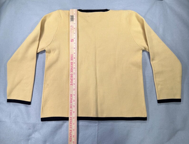 May include: A light yellow sweater with long sleeves and a crew neck. The sweater has black trim on the neckline, cuffs, and hem. A Home Depot ruler is placed vertically on the sweater, measuring its length.