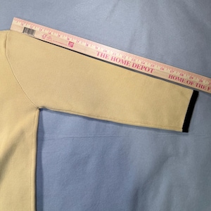 May include: A light yellow sweater sleeve with a black trim at the cuff. A wooden ruler is placed diagonally across the sleeve. The text "THE HOME DEPOT" and "HOME OF THE" is visible on the ruler.