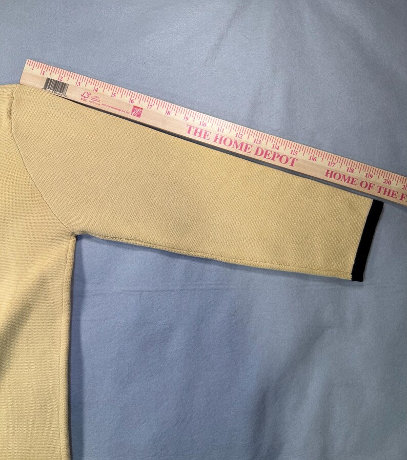 May include: A light yellow sweater sleeve with a black trim at the cuff. A wooden ruler is placed diagonally across the sleeve. The text "THE HOME DEPOT" and "HOME OF THE" is visible on the ruler.