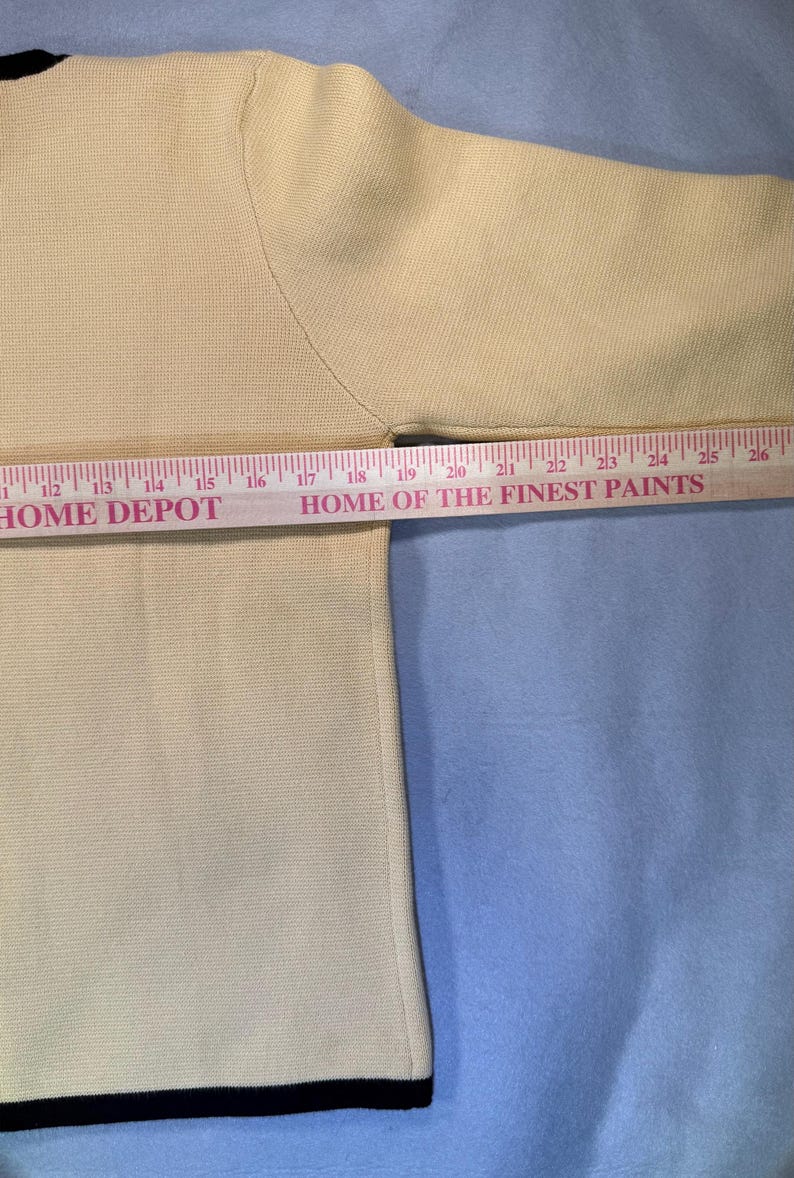 May include: A cream-colored sweater with a black trim at the bottom and neckline. The sleeve is visible, and a ruler is placed across the garment. The text "HOME DEPOT" and "HOME OF THE FINEST PAINTS" are visible.