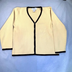May include: A cream-colored cardigan sweater with a V-neck and long sleeves. The sweater has black trim along the neckline, button placket, cuffs, and hem. It features a button-down front and a small label near the neckline.