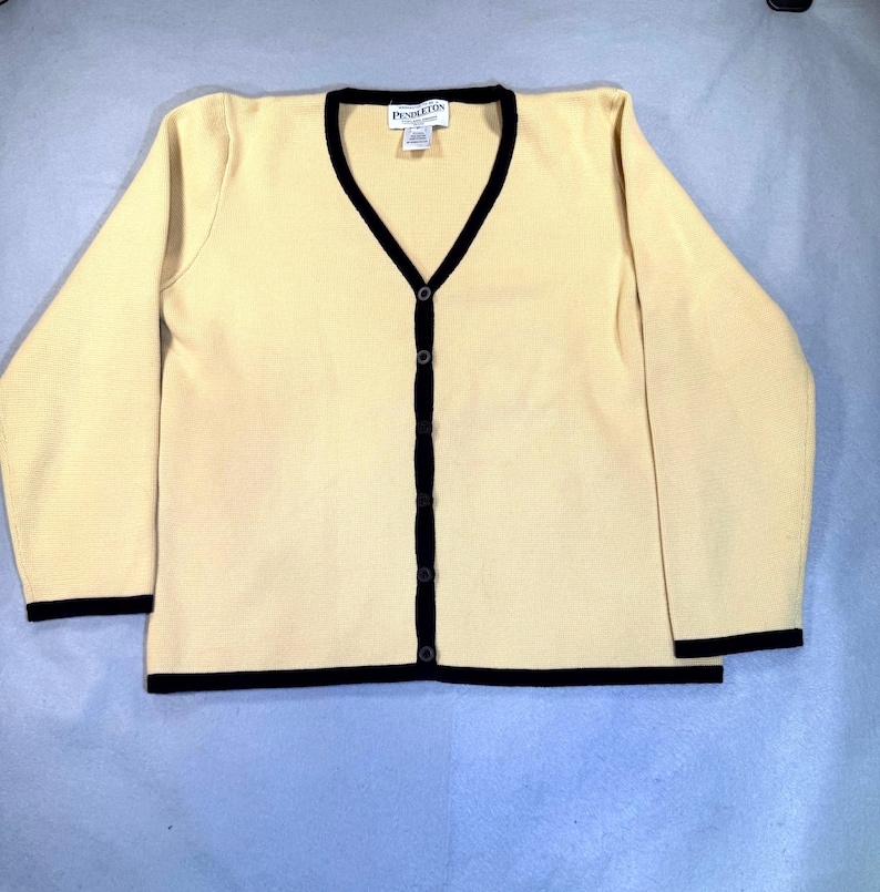 May include: A cream-colored cardigan sweater with a V-neck and long sleeves. The sweater has black trim along the neckline, button placket, cuffs, and hem. It features a button-down front and a small label near the neckline.