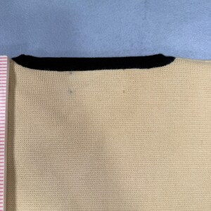 May include: Close-up of a cream-colored knit top with a black neckline. The fabric has a fine, textured weave. The top is set against a light blue background.
