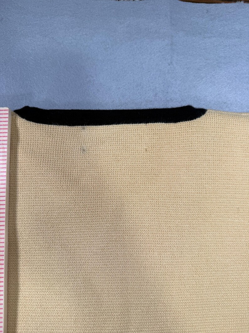 May include: Close-up of a cream-colored knit top with a black neckline. The fabric has a fine, textured weave. The top is set against a light blue background.