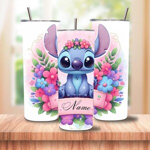 May include: Three white insulated tumblers with a cartoon character design. The central tumbler features a blue alien character surrounded by pink, purple, and blue flowers. The character has large eyes and is wearing a flower crown. The design includes the word "Name".