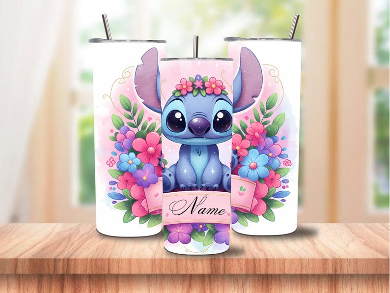 May include: Three white insulated tumblers with a cartoon character design. The central tumbler features a blue alien character surrounded by pink, purple, and blue flowers. The character has large eyes and is wearing a flower crown. The design includes the word "Name".