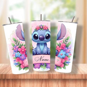 May include: A set of three white, insulated tumblers featuring a cartoon character with large ears and big eyes, surrounded by flowers. The character is blue with a flower crown. The tumblers have a pink and floral design with a straw.