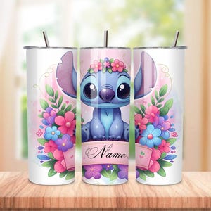 May include: A white tumbler featuring a blue cartoon character with large ears and a floral crown, surrounded by pink, purple, and blue flowers. The tumbler has a straw and a banner that says "Name".