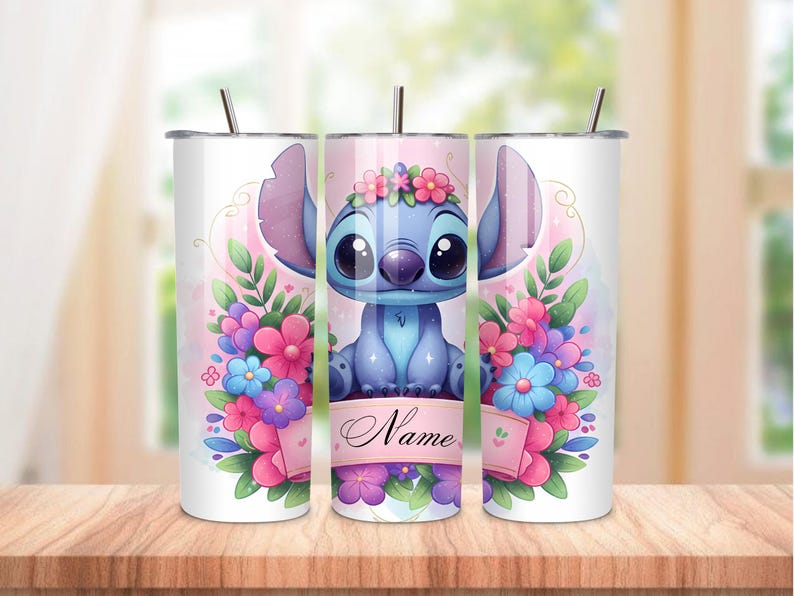 May include: A white tumbler featuring a blue cartoon character with large ears and a floral crown, surrounded by pink, purple, and blue flowers. The tumbler has a straw and a banner that says "Name".