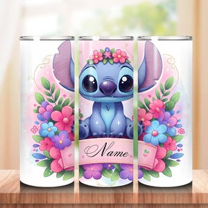 May include: A white tumbler featuring a cartoon character with large eyes, blue fur, and large ears, surrounded by colorful flowers. A pink banner with the word "Name" is across the bottom. The tumbler is on a wooden surface.