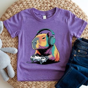 A purple t-shirt with a colorful cartoon image of a capybara wearing headphones and holding a video game controller.