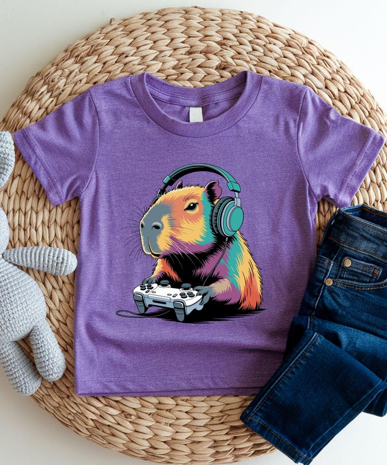 A purple t-shirt with a colorful cartoon image of a capybara wearing headphones and holding a video game controller.