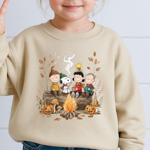 May include: A beige crewneck sweatshirt featuring a graphic print of the Peanuts characters sitting on a log by a campfire. The illustration includes Snoopy, Woodstock, Charlie Brown, Lucy, and Linus surrounded by autumn leaves, mushrooms, and pumpkins.