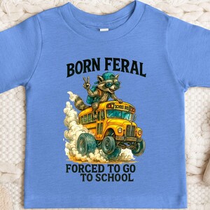 A blue t-shirt with a graphic design depicting a raccoon riding a school bus, with the text "BORN FERAL FORCED TO GO TO SCHOOL" printed on it.