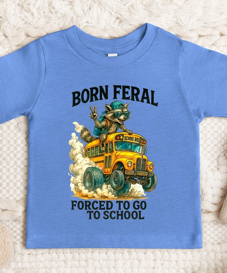 A blue t-shirt with a graphic design depicting a raccoon riding a school bus, with the text "BORN FERAL FORCED TO GO TO SCHOOL" printed on it.