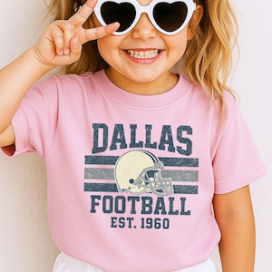 A young girl wearing a pink t-shirt with a graphic of a football helmet and the text "DALLAS FOOTBALL EST. 1960" is smiling and making a peace sign with her hand.