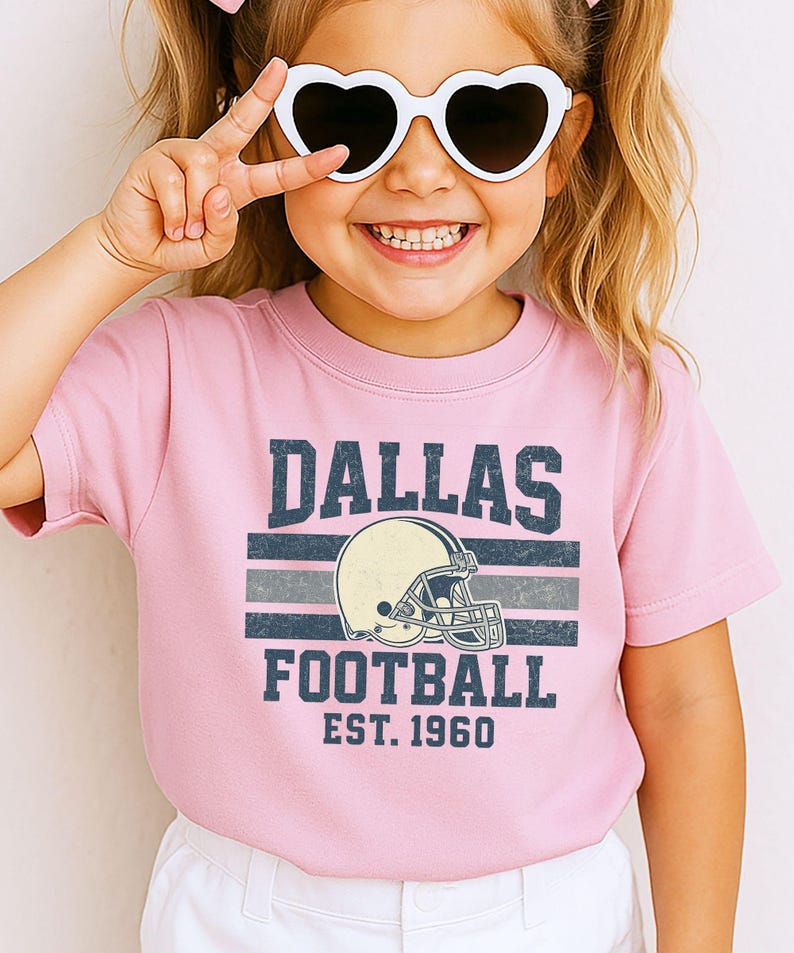 A young girl wearing a pink t-shirt with a graphic of a football helmet and the text "DALLAS FOOTBALL EST. 1960" is smiling and making a peace sign with her hand.