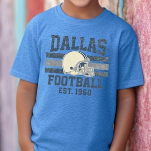 A young boy wearing a blue t-shirt with the text "DALLAS FOOTBALL EST. 1960" printed on it.