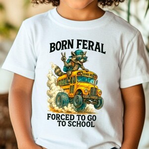 A young child wearing a white t-shirt with a graphic design of a raccoon riding a school bus.