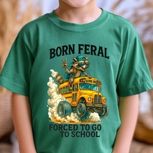 A young boy wearing a green t-shirt with a graphic design of a raccoon riding on the back of a school bus, along with the text "Born Feral Forced to Go to School".