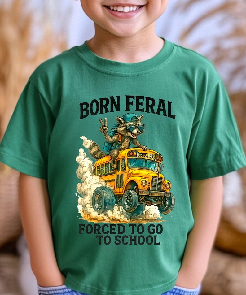 A young boy wearing a green t-shirt with a graphic design of a raccoon riding on the back of a school bus, along with the text "Born Feral Forced to Go to School".